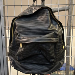 Svart ryggsäck från Mi-Pac - Snygg svart ryggsäck från Mi-Pac med ett coolt perforerat mönster. Väskan har guldfärgade dragkedjor och justerbara axelremmar. Perfekt för att bära dina grejer med stil! 🖤