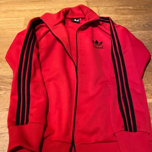 Röd vintage träningsjacka från Adidas - Snygg röd vintage original träningsjacka från Adidas med svarta ränder längs ärmarna. Jackan har dragkedja som och ribbade muddar vid ärmslut. Perfekt för en sportig look! Dragkedja är trasig men kan fixas av den händige! 