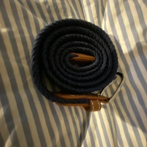 Blått flätat bälte med bruna detaljer - Snyggt blått flätat bälte med bruna läderdetaljer och silverfärgat spänne. Perfekt för att ge en stilren touch till din outfit. Passar både till jeans och chinos.