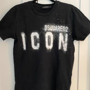 Svart t-shirt från Dsquared2 - Säljer en svart t-shirt från Dsquared2 med texten 'ICON' i vitt på framsidan. Den är kortärmad och i bra skick. Perfekt för en casual look eller att styla upp för en fest. Passar både till jeans och shorts! Hör av er vid minsta fundering och pris kan diskuteras👑