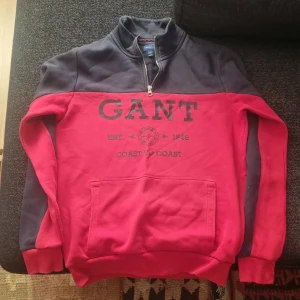Röd och marinblå tröja från GANT - Säljer en snygg och bekväm röd och marinblå tröja från GANT. Den har en dragkedja vid halsen och ett tryck med texten 'GANT EST. 1949 COAST TO COAST' på framsidan. Perfekt för en casual look och passar bra till både jeans och chinos. Tröjan är i bra skick och har anvädns sällan