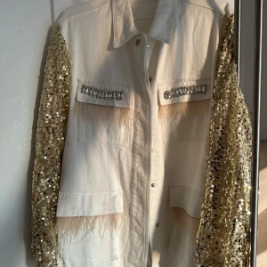 Vit jacka med paljettärmar - Säljer en unik vit jeansjacka med glittriga guldpaljettärmar. Cool design med fjäderdetaljer på framsidan och gnistrande stenar på fickorna. Perfekt för att sticka ut på festen! Inköpt på Raglady i våras och använd endast en gång. Superfin!