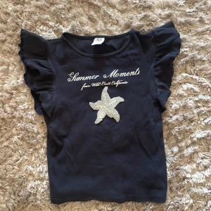 Svart topp med volangärmar från Zara - Säljer en snygg mörkblå topp från Zara med texten 'Summer Moments from West Coast California' och en glittrig sjöstjärna på framsidan. Toppen har volangärmar som ger en extra touch av stil. Perfekt för sommarens alla äventyr!