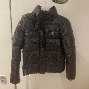 Svart pufferjacka från Calvin Klein Jeans - Säljer en snygg svart pufferjacka från Calvin Klein Jeans. Jackan är i storlek XS och har en glansig finish som ger en cool look. Den är perfekt för kalla dagar med sin vadderade design och har en praktisk huva. Jackan har långa ärmar och stängs med dragkedja och knappar framtill. Perfekt för vintern!