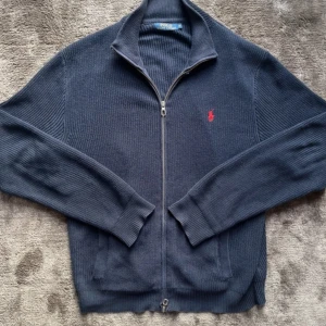 Mörkblå zip | Ralph Lauren  - Säljer en stilren mörkblå tröja från Polo Ralph Lauren med dragkedja och röd broderad logga på bröstet. Tröjan är ribbad och har långa ärmar, perfekt för kyligare dagar. 