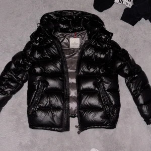 Svart pufferjacka från Moncler - Säljer en snygg svart pufferjacka från Moncler. Jackan har en glansig finish och är utrustad med dragkedja och två fickor framtill. Perfekt för kyliga dagar med sin vadderade design.