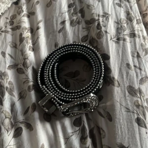 Svart glittrigt bälte med nitar - Snyggt svart bälte med glittriga detaljer och nitar. Bältet har en silverfärgad spänne dekorerad med strass. Perfekt för att ge en extra touch till din outfit.