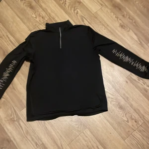 Svart långärmad träningströja från Crivit - Säljer en svart långärmad tröja från Crivit med dragkedja vid halsen. Perfekt för kyligare dagar och har en stilren design med ett diskret mönster på ärmen. Tröjan är tillverkad i ett mjukt och bekvämt material.