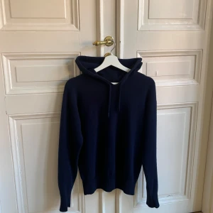 Mörkblå hoodie från Carmes - Säljer en stilren mörkblå hoodie från Carmes i storlek S. Tröjan är tillverkad i 100% kashmir vilket gör den extra mjuk och bekväm. Perfekt för kyliga dagar med sin klassiska design och praktiska huva.