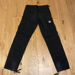 Svarta cargopants från Carhartt - Säljer ett par svarta cargopants från Carhartt. Byxorna har flera praktiska fickor och justerbara band vid bensluten. Perfekta för en avslappnad och funktionell stil.