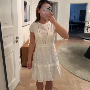 Vit klänning med spetsdetaljer - Charmig vit klänning med spetsdetaljer och korta ärmar. Den har en öppen rygg med snören vid nacken. Perfekt för sommarens alla tillfällen. Använd endast en gång. 