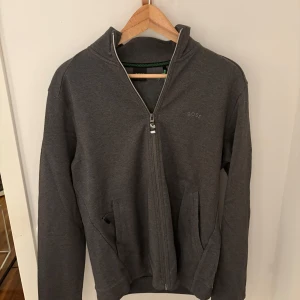 Grå jacka från Boss - Snygg grå jacka från Boss med dragkedja och två fickor framtill. Perfekt för en stilren look. Jackan har en diskret logga på bröstet och är tillverkad i ett mjukt material. Storlek Medium. Pris går att diskutera om ni väcker intresse. 