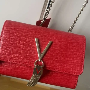 Röd axelväska med kedja - Snygg röd axelväska från Valentino med en elegant kedjerem och metallisk V-detalj framtill. Väskan har en strukturerad yta och stängs med en tryckknapp. Perfekt för att ge en färgklick till din outfit. Den är aldrig använd och har fortfarande kvar prislapp. Skriv vid intresse eller frågor❤️