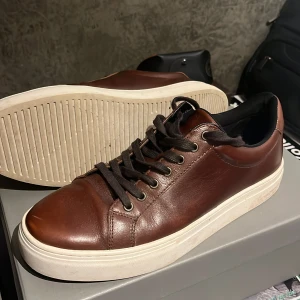 Bruna sneakers från Vagabond - Snygga bruna sneakers från Vagabond i skinn med svarta snören och vit sula. Perfekta för en stilren look. Skorna har en klassisk design med rund tå och snörning. Storlek är 41 passar typ 42. Skick är 9/10 knappt använda bara lite cresade men de går att fräscha upp. Nypris runt 1400:-