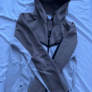 Nike tech fleece - Säljer en snygg tech fleece från Nike i beige och vitt. Tröjan har en dragkedja framtill och en justerbar huva. Perfekt för en avslappnad stil. Loggan är lite sliten men utöver det i bra skick. Storlek S