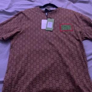 Beige t-shirt från Gucci - Säljer en beige t-shirt från Gucci med det ikoniska GG-mönstret över hela tyget. T-shirten har en grön och röd logotyp på bröstet och är kortärmad. Perfekt för den som vill ha en stilren och lyxig look. 8000 kroner ny pris. Aldrig använd prislappen sitter på