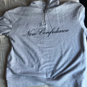 Grå hoodie med text och fjärilar - Säljer en grå hoodie med texten 'New Confidence' på framsidan och tre fjärilar på ärmen. Tröjan har en dragkedja vid halsen och ribbade muddar.