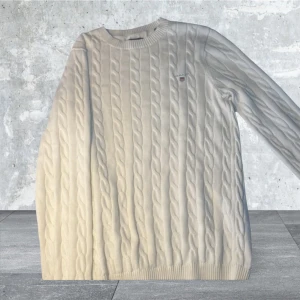 Vit kabelstickad tröja från Gant - Säljer en stilren vit kabelstickad tröja från Gant. Tröjan har en klassisk design med rund hals och långa ärmar. Perfekt för en elegant och avslappnad look. 🧶✨( Helt oanvänd )!