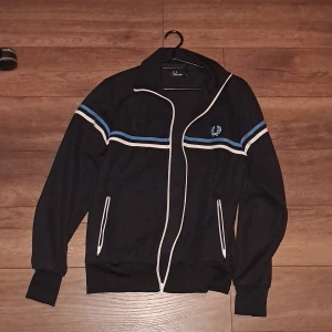 Svart träningsjacka från Fred Perry - Säljer en svart träningsjacka från Fred Perry med vita och blå ränder. Jackan har dragkedja och långa ärmar, samt ribbade muddar vid ärmslut och nederkant. Perfekt för en sportig look! 