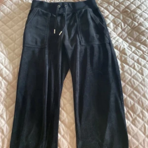 Svarta byxor från Juicy Couture - Snygga svarta byxor från Juicy Couture med dragsko i midjan och praktiska fickor. Byxorna har en stilren design med en liten metallknappdetalj på fickan. Perfekta för en avslappnad och trendig look.