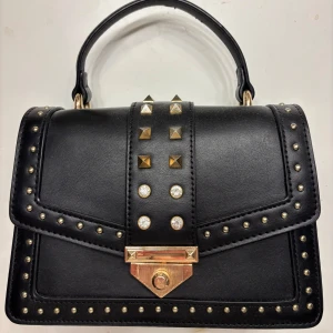 Svart handväska med nitar - Snygg svart handväska med guldfärgade nitar och detaljer. Väskan har ett handtag och en avtagbar axelrem. Den stängs med ett elegant guldfärgat lås och har flera fack inuti för enkel organisering. Perfekt för att ge din outfit en edgy touch.