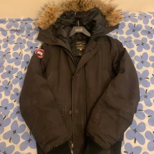 Svart parkajacka från Canada Goose - Säljer en svart parkajacka från Canada Goose med pälsfodrad huva och ikonisk logga på ärmen. Jackan har dragkedja och knappar framtill samt flera praktiska fickor. Perfekt för kalla vinterdagar. Är 188 och den passar mig perfekt så skulle säga att den är s inte 2xs