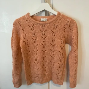 Persiko färgad  stickad tröja från Name It - Säljer en söt rosa stickad tröja från Name It med ett fint hålmönster. Tröjan har långa ärmar och rund halsringning, perfekt för kyligare dagar. Passar barn i åldern 9-10 år.