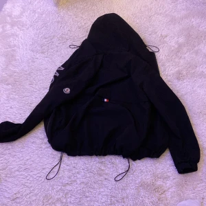 Svart vindjacka från Moncler - Säljer en stilren svart vindjacka från Moncler med huva och dragkedja. Jackan har långa ärmar och justerbara snören i nederkant för en perfekt passform. Moncler-logga på ärmen och stort tryck på framsidan. Perfekt för blåsiga dagar!