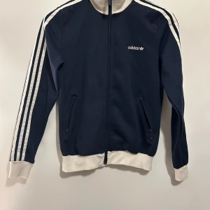 Träningströja adidas - Klassisk Adidas Originals träningströja tre ränder längs ärmarna och Adidas-loggan broderad på bröstet. Jackan har en krage och muddar i ribbstickat material, samt dragkedja fram och fickor med dragkedja. Bra skick, storlek xs/s