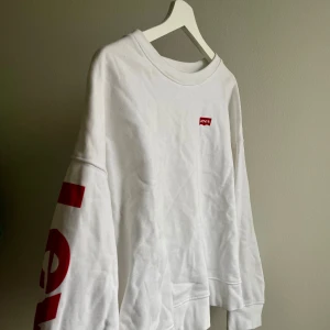 Vit långärmad tröja från Levi's - Säljer en vit långärmad tröja från Levi's med röd logga på bröstet och tryck på ärmen. Perfekt för en avslappnad stil. Tröjan har en rund halsringning och är gjord i mjukt material. Använd fåtal gånger, lite boxigare i modellen.