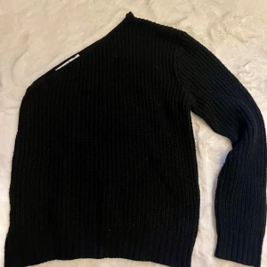 Svart stickad tröja från NA-KD - Snygg svart stickad tröja från NA-KD med en unik oneshoulder-design. Perfekt för att ge din outfit en trendig touch. Tröjan har långa ärmar och en ribbad struktur som ger en skön passform. Använd fåtal gånger!