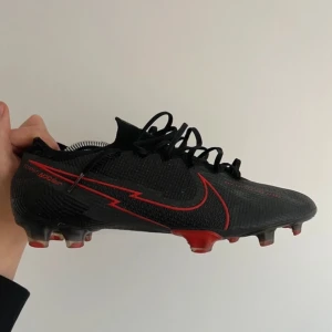 Nike mercurial vapor  - Säljer ett par svarta Nike fotbollsskor med röda detaljer och snörning. Skorna har en modern design med en tydlig Nike-logga i rött på sidan. Perfekta för fotbollsplanen med sina greppvänliga dobbar.