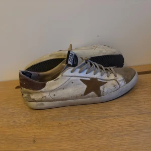 Sneakers från Golden Goose - Säljer ett par vita sneakers från Golden Goose med bruna detaljer och en stjärna på sidan. Skorna har snörning och en sliten look som är typisk för märket. Perfekta för en avslappnad stil. Storlek 43. PRIS KAN DISKUTERAS!