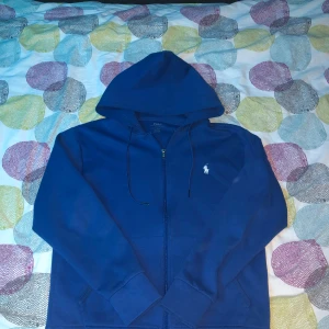 Blå hoodie från Polo Ralph Lauren - Säljer en snygg blå hoodie från Polo Ralph Lauren med dragkedja och det klassiska logotypbroderiet på bröstet. Perfekt för en avslappnad stil. Den har långa ärmar och en praktisk huva med dragsko.obs har inget kvitto pgr av slarv så hittar ja inte de,, tar imot byte