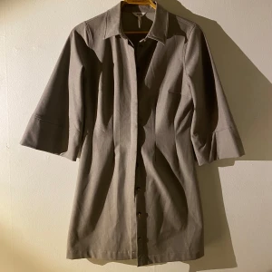 Beige överdel med trekvartsärm - Snygg beige kappa med trekvartsärm och knappar framtill. Perfekt för vår och höst med sin stilrena design och bekväma passform. En tidlös klassiker för garderoben.