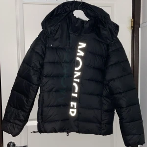 Svart dunjacka från Moncler - Säljer en snygg svart dunjacka från Moncler med stor logotyp på framsidan. Jackan har en avtagbar huva och dragkedja. Perfekt för kalla dagar med sin quiltade design. OBS! Priset kan diskuteras