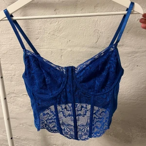 Blå spetskorsett - Snygg blå korsett med spetsdetaljer och justerbara axelband. Perfekt för en stilfull look med en touch av elegans. Den har en vacker spetsdesign framtill och en slät baksida.