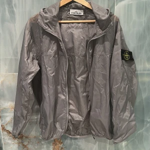 Grå vindjacka från Stone Island - Säljer en stilren grå vindjacka från Stone Island. Jackan har en dragkedja framtill och en praktisk huva. Den ikoniska logotypen finns på ärmen. Perfekt för blåsiga dagar.