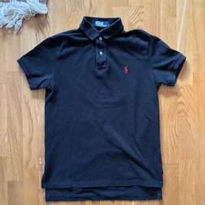 Svart pikétröja från Ralph Lauren - Säljer en klassisk svart pikétröja från Ralph Lauren med röd broderad logga på bröstet. Tröjan har korta ärmar och knappar vid kragen. Perfekt för en stilren look.