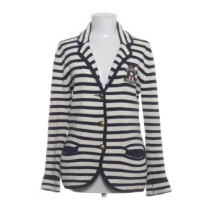 Randig kavaj från Lexington - Säljer en stilren randig kavaj från Lexington i blått och vitt. Kavajen har långa ärmar, två knappar framtill och ett broderat emblem på bröstet. Tillverkad i 100% bomull för en bekväm känsla. Perfekt för en klassisk look!