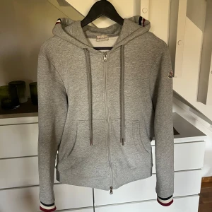 Grå zip hoodie moncler  - Säljer en stilren grå hoodie från Moncler med dragkedja och huva. Tröjan har ett broderat Moncler-märke på ärmen och på luvan och längst ner på ärmarna syns monclers färger, för mer bilder eller frågor är de bara att skriva kvitto finns!