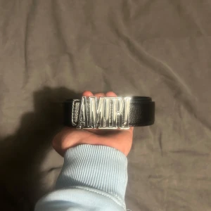 Svart bälte med AMIRI-spänne - Snyggt svart bälte från AMIRI med ett iögonfallande silverfärgat spänne som stavar ut märkesnamnet. Perfekt för att ge en stilren touch till din outfit.Köpte den när jag var i Italien. 