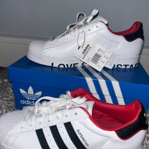 Adidas Superstar sneakers - Snygga Adidas Superstar sneakers i vitt med svarta ränder och röd insida. Skorna har klassisk snörning och textdetaljer på sulan. Perfekta för en stilren look. Sprillans nya med taggen kvar, köpte dem och glömde bort dem. 