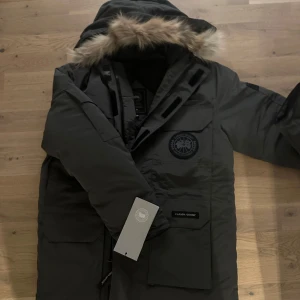 Canada goose - Säljer en grå parkajacka från Canada Goose med avtagbar pälsdetalj på huvan. Jackan har en långärmad design och är perfekt för kalla vinterdagar. Den har flera praktiska fickor och en dragkedja med kardborreknäppning för extra skydd mot kyla.