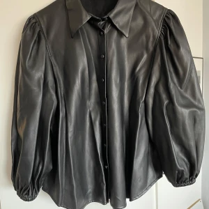Svart skinnskjorta från Zara - Säljer en snygg svart skinnskjorta från Zara. Den har en cool oversized passform med långa puffärmar och knappar framtill. Perfekt för en stilren look, oavsett om det är till vardags eller fest. XL låter stort men den är inte så stor