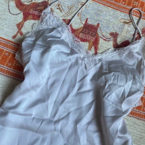 Vit sidenklänning med spetsdetaljer - Säljer en elegant silver/vit klänning i siden med vackra spetsdetaljer längs halsringningen. Klänningen har  justerbara axelband och har en mjuk och lyxig känsla. Perfekt för sommarens fester, en romantisk kväll eller lyxigt nattlinne. 💫