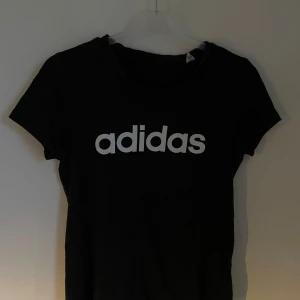 Svart t-shirt från Adidas - Säljer en svart t-shirt från Adidas med deras klassiska logga i vitt på bröstet. T-shirten är kortärmad och perfekt för en avslappnad stil. Passar både till träning och vardags.