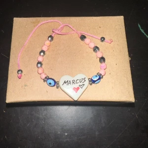 Armband med hjärta och pärlor - Säljer ett supergulligt armband med ett hjärta i mitten där det står 'Marcus' och en liten röd hjärta bredvid. Armbandet har rosa och svarta pärlor samt blå ögonpärlor. Det är justerbart med ett rosa snöre. Perfekt för att ge bort som en personlig present eller bara för att bära själv!