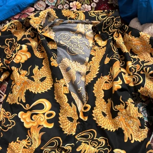 Svart och guldmönstrad skjorta med drakar - Säljer en cool svart skjorta med ett iögonfallande guldmönster av drakar. Skjortan har långa ärmar och knappar framtill. Perfekt för att sticka ut på festen eller andra speciella tillfällen. Materialet känns mjukt och bekvämt. Passar perfekt för dig som gillar unika och djärva stilar!