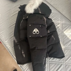 Svart pufferjacka med pälsdetalj - Säljer en snygg svart pufferjacka från Moose Knuckles. Den har en fluffig vit pälsdetalj på huvan och en cool logga på ärmen. Perfekt för kalla vinterdagar! Jackan har dragkedja och knappar för extra skydd mot kylan. Passar både till vardags och fest. 🖤